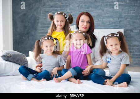 Large friendly family, many children: mom and four pretty cheerful girls triple twins sisters sitting on a bed against a gray brick wall. Stock Photo