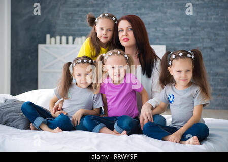 Large friendly family, many children: mom and four pretty cheerful girls triple twins sisters sitting on a bed against a gray brick wall. Stock Photo