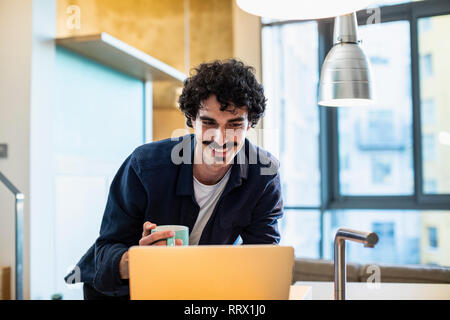 Smiling man drinking coffee, working at laptop in apartment kitchen Stock Photo