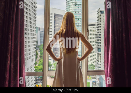 Young woman opens the window curtains and looks at the skyscrapers in the big city Stock Photo