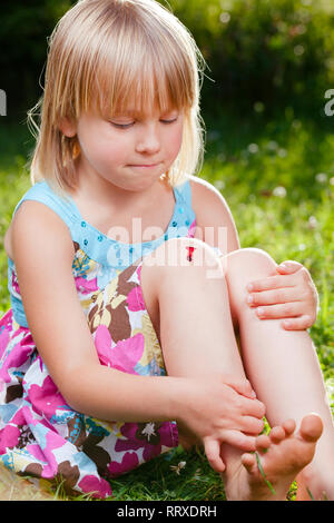 Little girl wearing summer dress sitting on a lawn in a garden with small injury on her knee. She is looking at aching place with pursed lips holding  Stock Photo