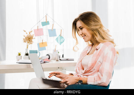 Smiling blonde woman in checkered shirt typing on laptop keyboard Stock Photo
