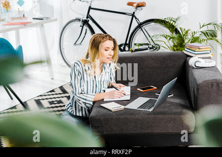 Woman in striped shirt writing in notebook and looking at laptop Stock Photo