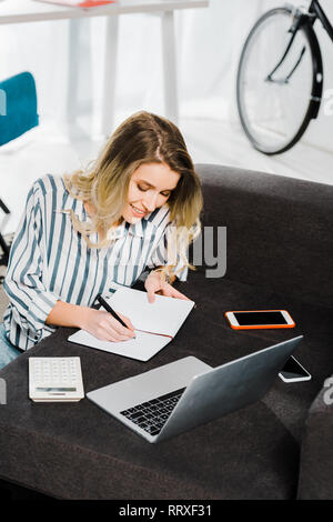 Smiling woman with laptop and smartphones writing in notebook Stock Photo