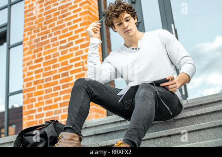 Attractive man holding notepad in his hands and sitting on concrete steps of red bricked building. Stock Photo
