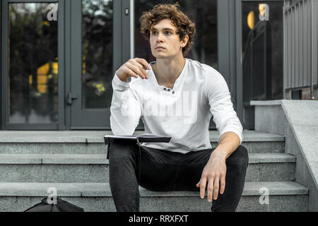 Attractive pensive student sitting on modern building steps and making notes in his black notepad. Stock Photo