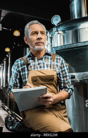 thoughtful brewer in working overalls writing in notepad near brewery equipment Stock Photo