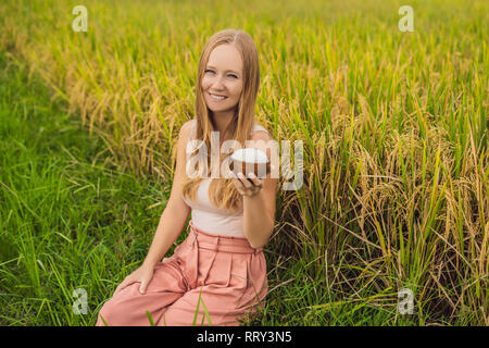 A woman is holding a cup of boiled rice in a wooden cup on the background of a ripe rice field Stock Photo