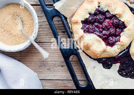 Homemade cherry galette on backing tray on wooden table with brown sugar. Process of baking. Open pie, cherries tart. Berries bakery. Summer pies on p Stock Photo