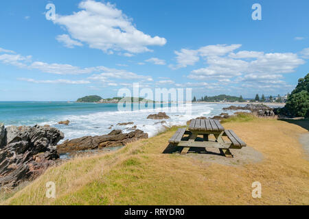 Outdoor table beside walking track with distant waves smoothed by long exposure washing in around rocky foreshore at base of Mount Maunganui, Tauranga Stock Photo