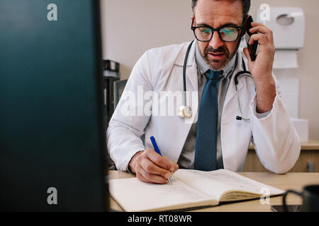 Mature male doctor talking on cell phone and making notes in his book. Physician making phone call and writing in his diary. Stock Photo