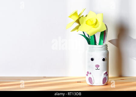 DIY Easter vase bunny with narcissus, daffodils from glass jar, felt, googly eyes on a white wall background. Gift ideas, decor for spring, Easter. Ha Stock Photo