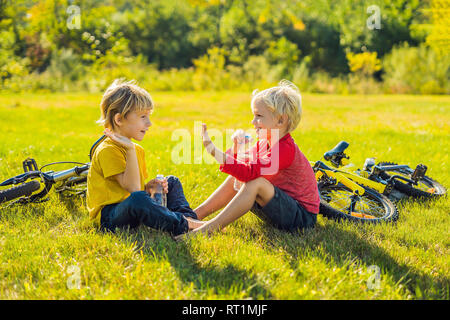 Two little boys drink water in the park after riding a bike Stock Photo