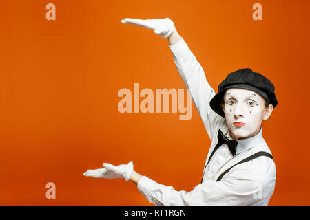 Emotional pantomime with white facial makeup showing empty space on the orange background, advertising something Stock Photo
