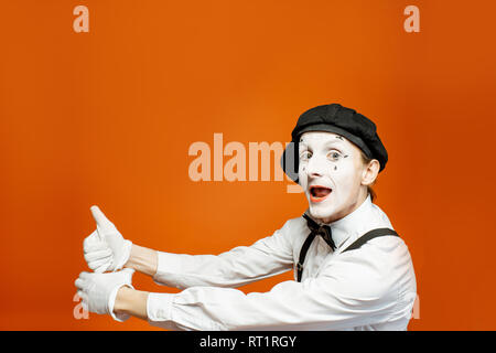 Emotional pantomime with white facial makeup showing empty space on the orange background, advertising something Stock Photo