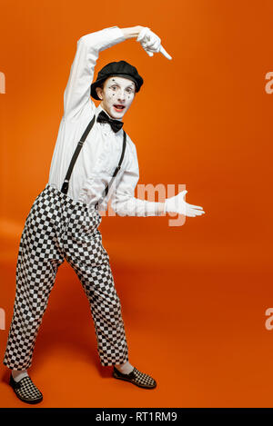 Emotional pantomime with white facial makeup showing empty space on the orange background, advertising something Stock Photo