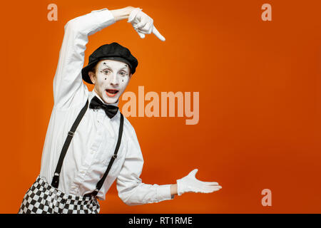 Emotional pantomime with white facial makeup showing empty space on the orange background, advertising something Stock Photo