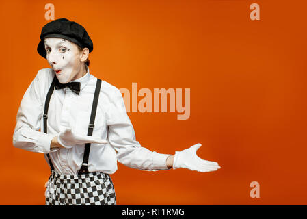 Emotional pantomime with white facial makeup showing empty space on the orange background, advertising something Stock Photo