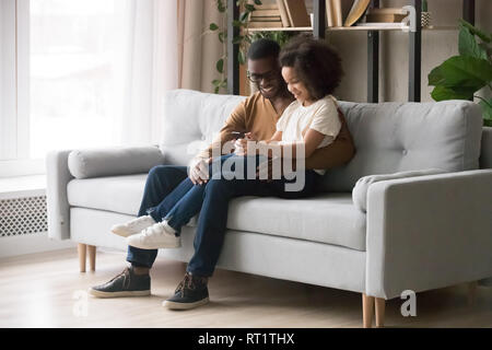 Happy kid girl sitting on african dad lap holding phone Stock Photo