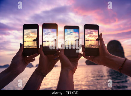 Closeup photo of four hands with mobile phones taking photo of beautiful sunset over sea bay Stock Photo