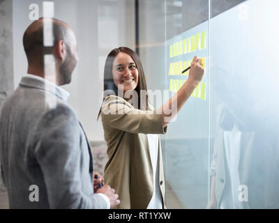 Businesswoman smiling at colleague writing on sticky notes at glass pane in office Stock Photo