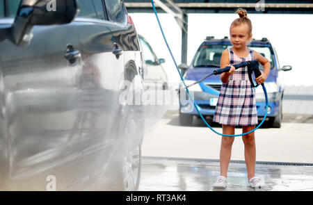 Pretty serious little preschool girl in summer dress helping to parents use coin-operated self-serve car wash, clean auto Stock Photo