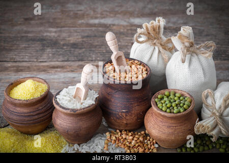 Rustic clay pots filled with rice, green mung, corn, buckwheat and sacks of grain on wooden table. Stock Photo