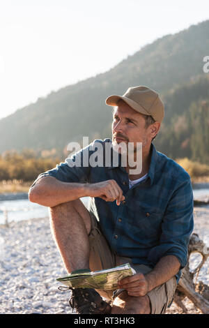 Mature man camping at riverside, sitting on tree trunk with map Stock Photo