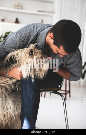 Young man sitting on chair cuddling dog Stock Photo