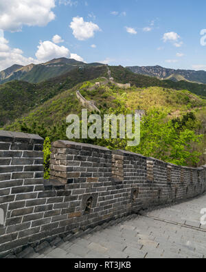 The steep stone steps of The Great Wall Of China at Mutianyu in the foreground. Several watchtowers can be seen as the wall climbs over the mountains. Stock Photo