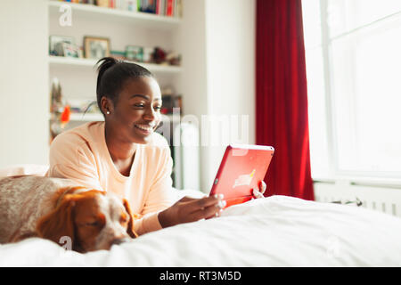 Smiling young woman using digital tablet next to sleeping dog on bed Stock Photo