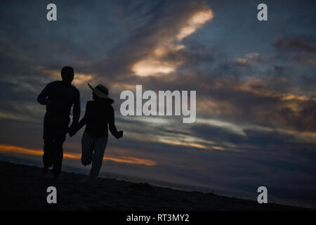 Smiling mid adult couple holding hands and running along a remote beach at sunset. Stock Photo