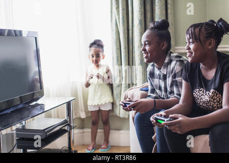 Toddler girl watching tween sisters playing video game in living room Stock Photo