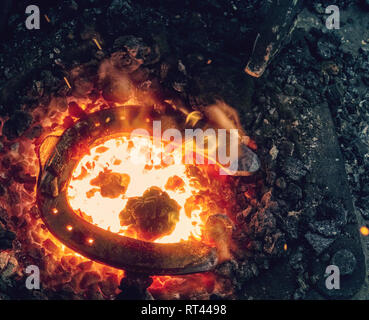 Red fire blazes inside of coal forge.  Blacksmith traditional works with hot metal  to form horse shoe from semiproduct Stock Photo