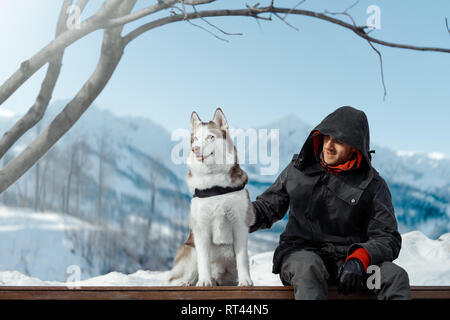 Man and Cute Siberian Husky dog sitting on bench highly in mountains on sunny winter day. Concept of animal wellbeing and owner care and love. Stock Photo