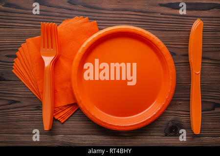 flat lay of stacked orange disposable plastic knives, forks and empty plates on napkins over wooden background Stock Photo