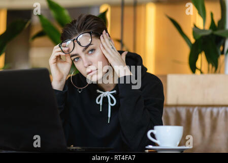 Portrait of tired shortcut hipster girl sitting with laptop at the cafe interior Stock Photo