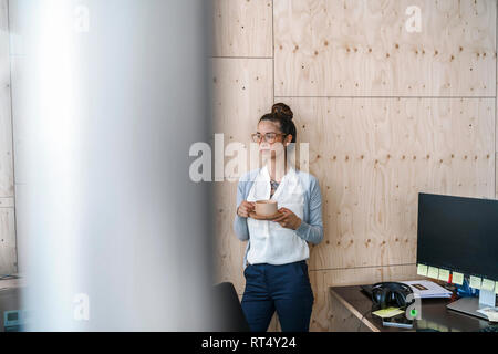 Young woman working in creative office, taking a break, drinking coffee from wooden cup