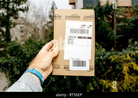 PARIS, FRANCE - NOV 28, 2017: Male hand holding the Amazon cardboard box as he picked it from the front door with trees and houses in the background  Stock Photo