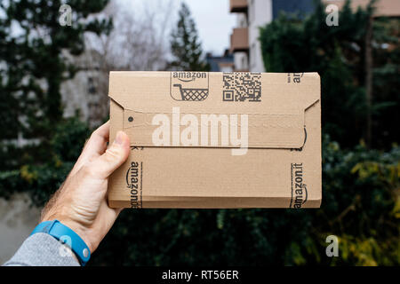 PARIS, FRANCE - NOV 28, 2017: Male hand holding the Amazon cardboard box as he picked it from the front door with trees and houses in the background  Stock Photo