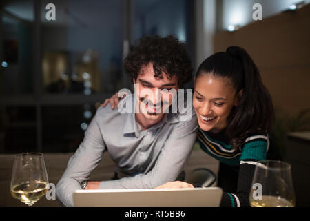 Happy couple using laptop and drinking white wine at home at night Stock Photo