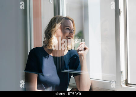 Happy blond woman in sunlight looking out of window Stock Photo