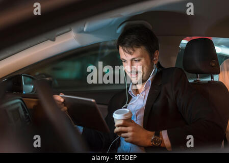 Businessman sitting in car at night, using digital tablet, drinking coffee Stock Photo