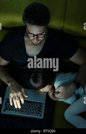 Father using laptop on couch at night with daughter  lying next to him Stock Photo