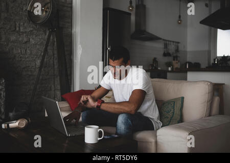 Young man sitting on sofa at home using laptop and checking the time Stock Photo