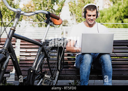 Young man sitting on a bench wearing headphones and using laptop next to bicycle Stock Photo