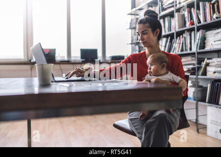 Working mother with baby on her lap, sitting in office, using laptop Stock Photo