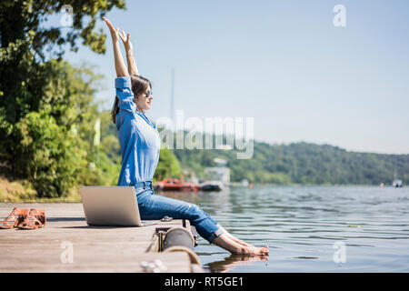 Woman with laptop sitting on a jetty at a lake, stretching Stock Photo