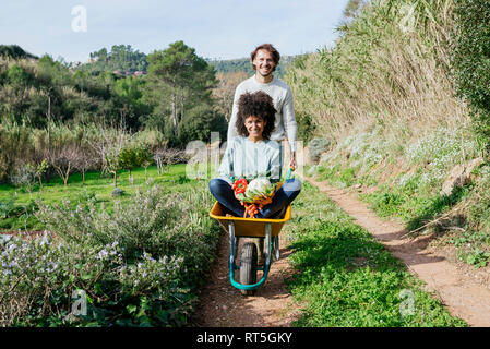 Woman sitting in wheelbarrow, holding fresh vegetables, man pushing her Stock Photo