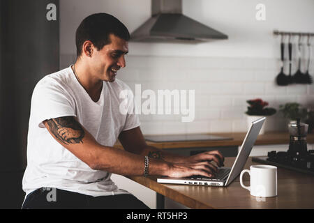 Smiling young man using laptop in kitchen at home Stock Photo